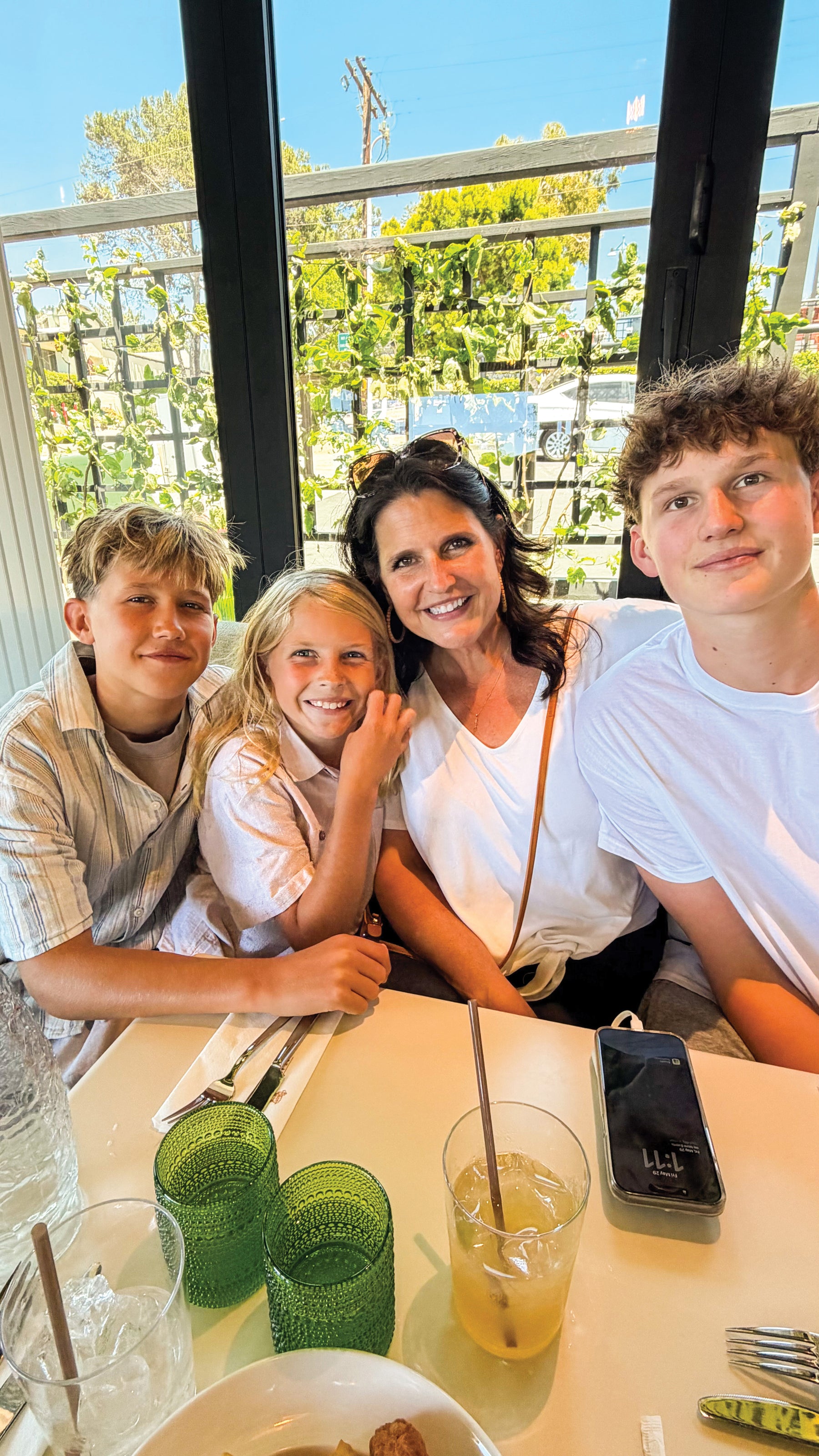 A family of four smiling and posing together at a restaurant table, with a woman in the center wearing a white top and a man on the right in a white t-shirt. The table has green textured glasses, a drink, and a smartphone, with a bright window background showing greenery outside.