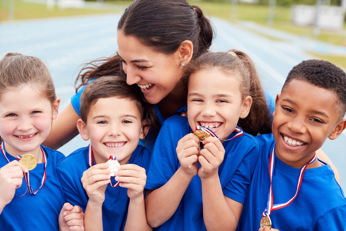 Five children wearing matching blue athletic shirts and medals hugging on an outdoor track, smiling and holding their medals.