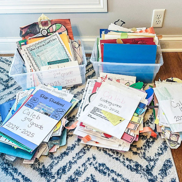 Stacks of school papers and folders on a carpeted floor with plastic bins.